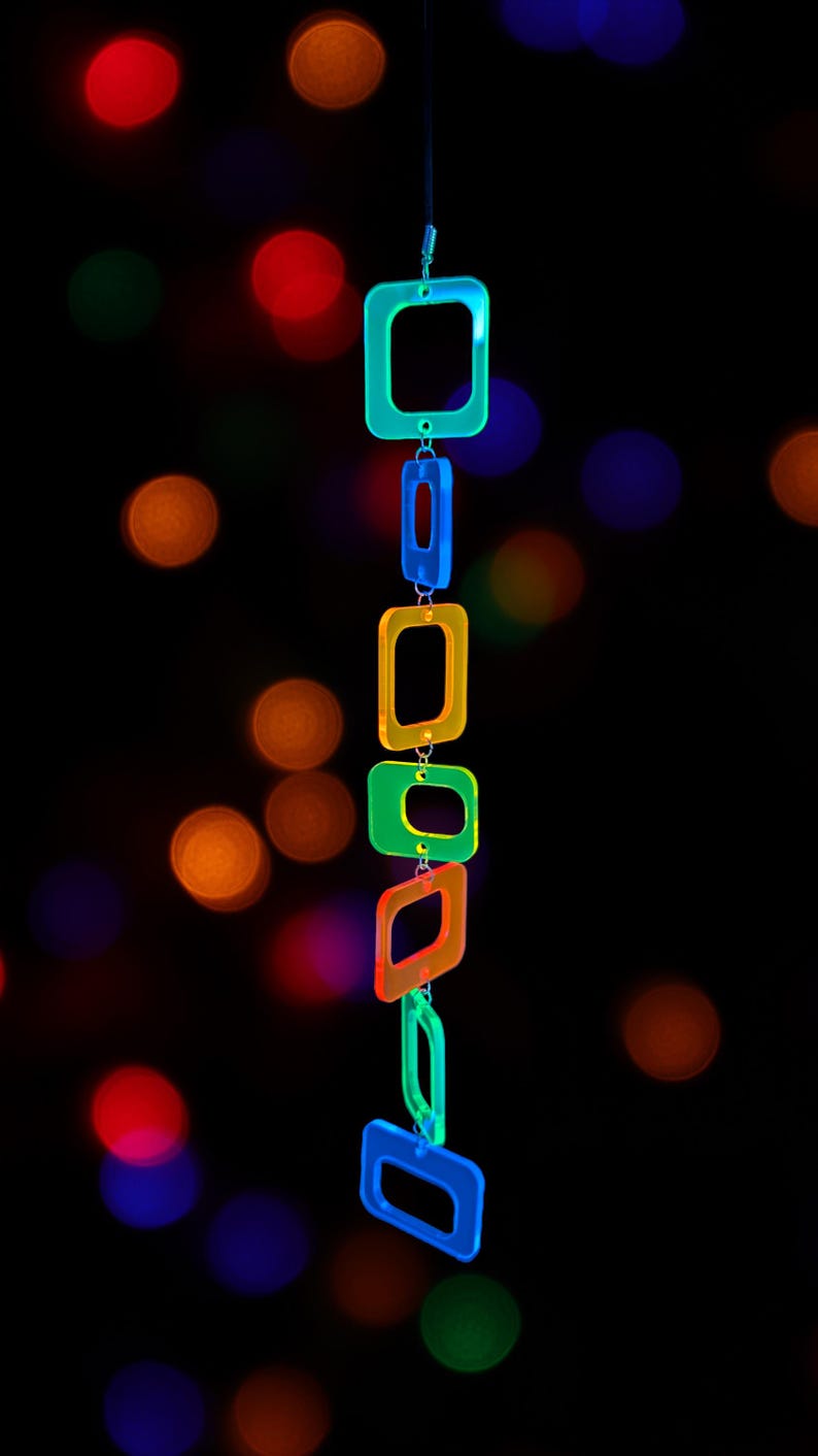 May include: A vertical hanging mobile composed of six translucent, neon-coloured square frames linked together. The frames are arranged in a sequence of light blue, dark blue, yellow, green, orange, and light blue, suspended by a thin black string.