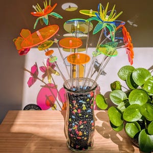 May include: A collection of translucent acrylic flower sculptures in vibrant neon orange, yellow, green, and blue hues. Each flower is mounted on a clear acrylic stem and arranged in a clear glass vase filled with small, multicolored decorative stones.