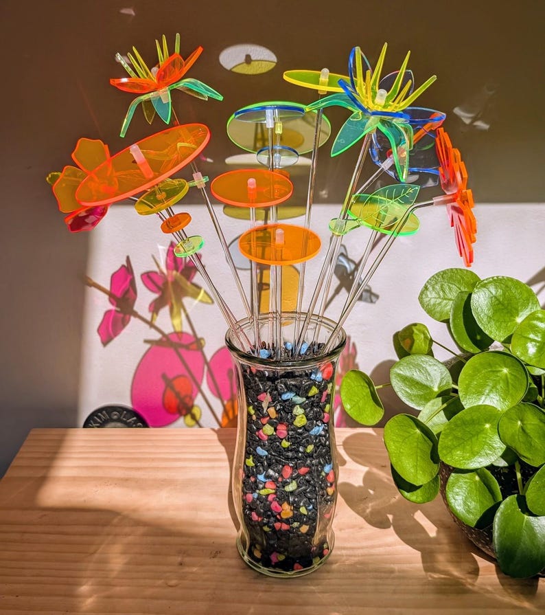 May include: A collection of translucent acrylic flower sculptures in vibrant neon orange, yellow, green, and blue hues. Each flower is mounted on a clear acrylic stem and arranged in a clear glass vase filled with small, multicolored decorative stones.