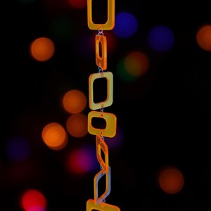 May include: A vertical hanging chain composed of translucent orange and yellow square-shaped links. The links are connected by small silver-toned metal jump rings, creating a flexible, geometric structure that hangs straight down against a dark background.