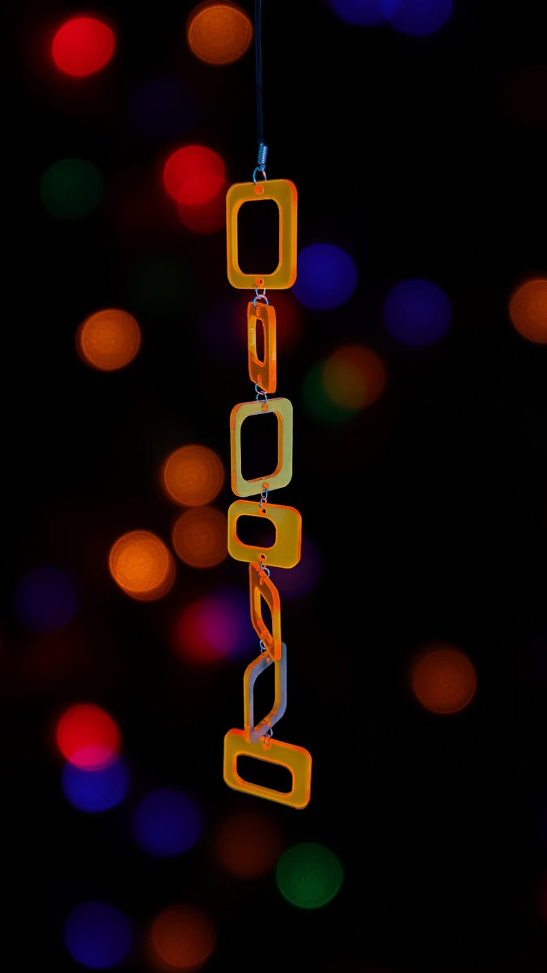 May include: A vertical hanging chain composed of translucent orange and yellow square-shaped links. The links are connected by small silver-toned metal jump rings, creating a flexible, geometric structure that hangs straight down against a dark background.