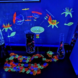 May include: A collection of neon-colored, UV-reactive plastic floral and geometric shapes arranged in clear glass vases. The set includes various flower-like structures, circular discs, and a scattered pile of small, colorful rectangular and square tiles.