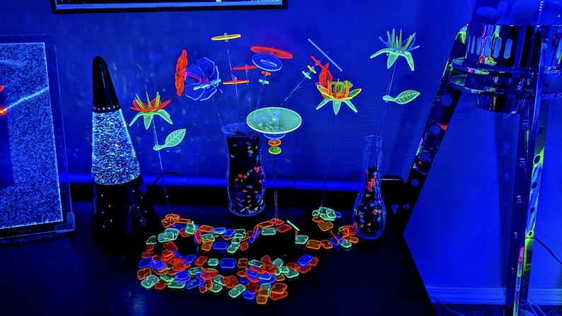 May include: A collection of neon-colored, UV-reactive plastic floral and geometric shapes arranged in clear glass vases. The set includes various flower-like structures, circular discs, and a scattered pile of small, colorful rectangular and square tiles.