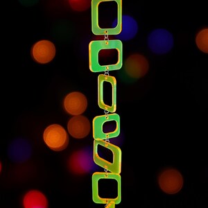 May include: A vertical chain of six translucent neon yellow-green square and rectangular acrylic links connected by small silver metal jump rings. The links are arranged in a descending line, creating a geometric pendant structure with a bright glow.
