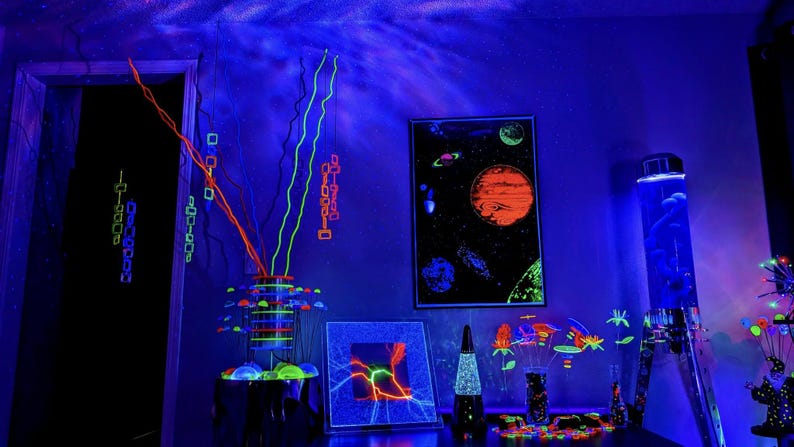 May include: A room interior illuminated by blue light featuring a space-themed poster, a lava lamp, a plasma disc, and several decorative arrangements of neon-coloured, glowing branches and floral shapes that emit a bright fluorescent light.