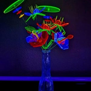 May include: A clear glass vase containing a bouquet of neon-colored plastic flowers that glow under ultraviolet light. The arrangement features vibrant red, blue, and green petals with thin stems, accented by circular green discs and small colorful beads.