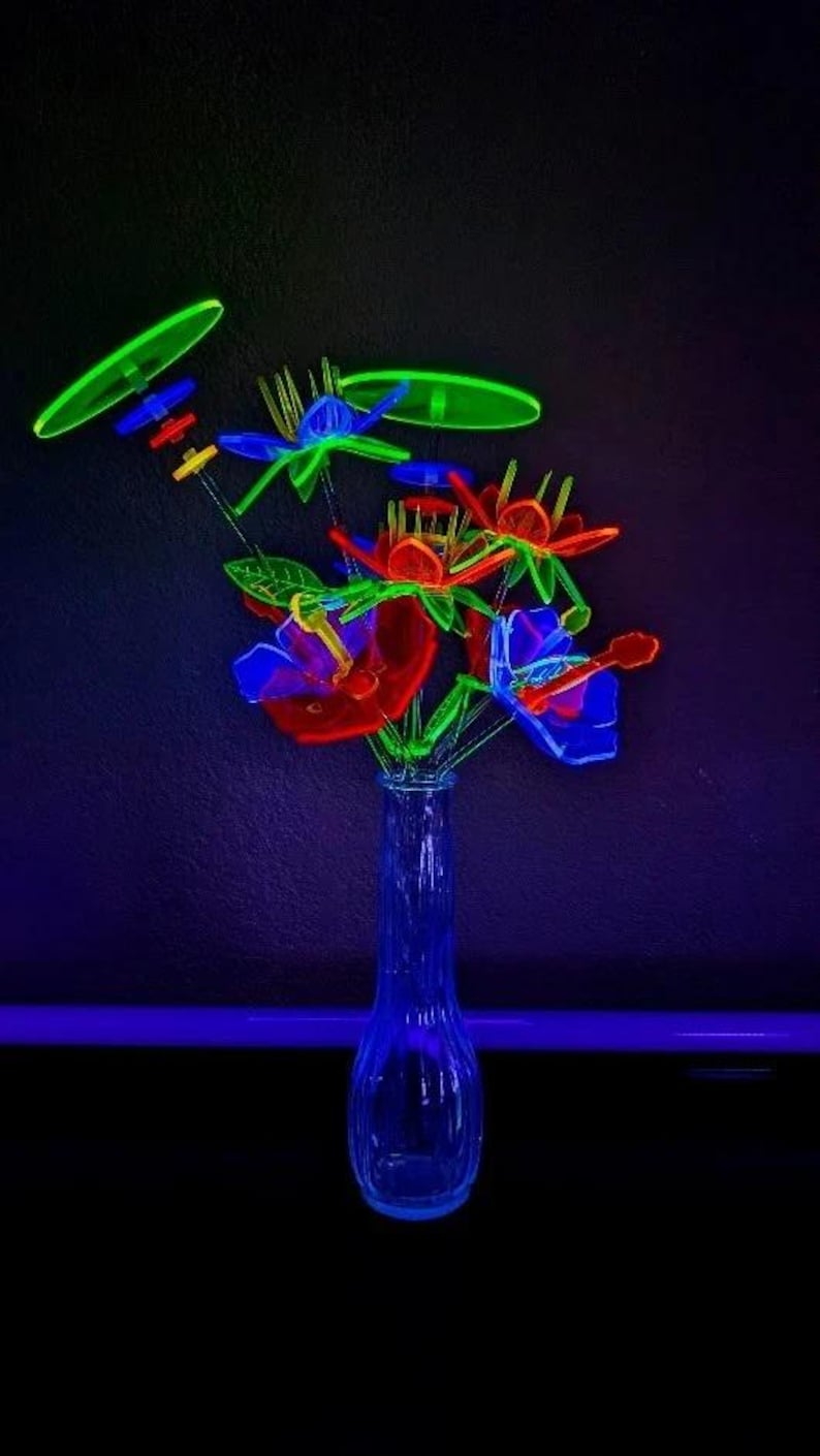 May include: A clear glass vase containing a bouquet of neon-colored plastic flowers that glow under ultraviolet light. The arrangement features vibrant red, blue, and green petals with thin stems, accented by circular green discs and small colorful beads.