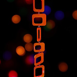 May include: A vertical hanging ornament composed of six translucent neon orange acrylic square and rectangular frames linked together by small silver metal jump rings, suspended from a thin black cord against a dark background.