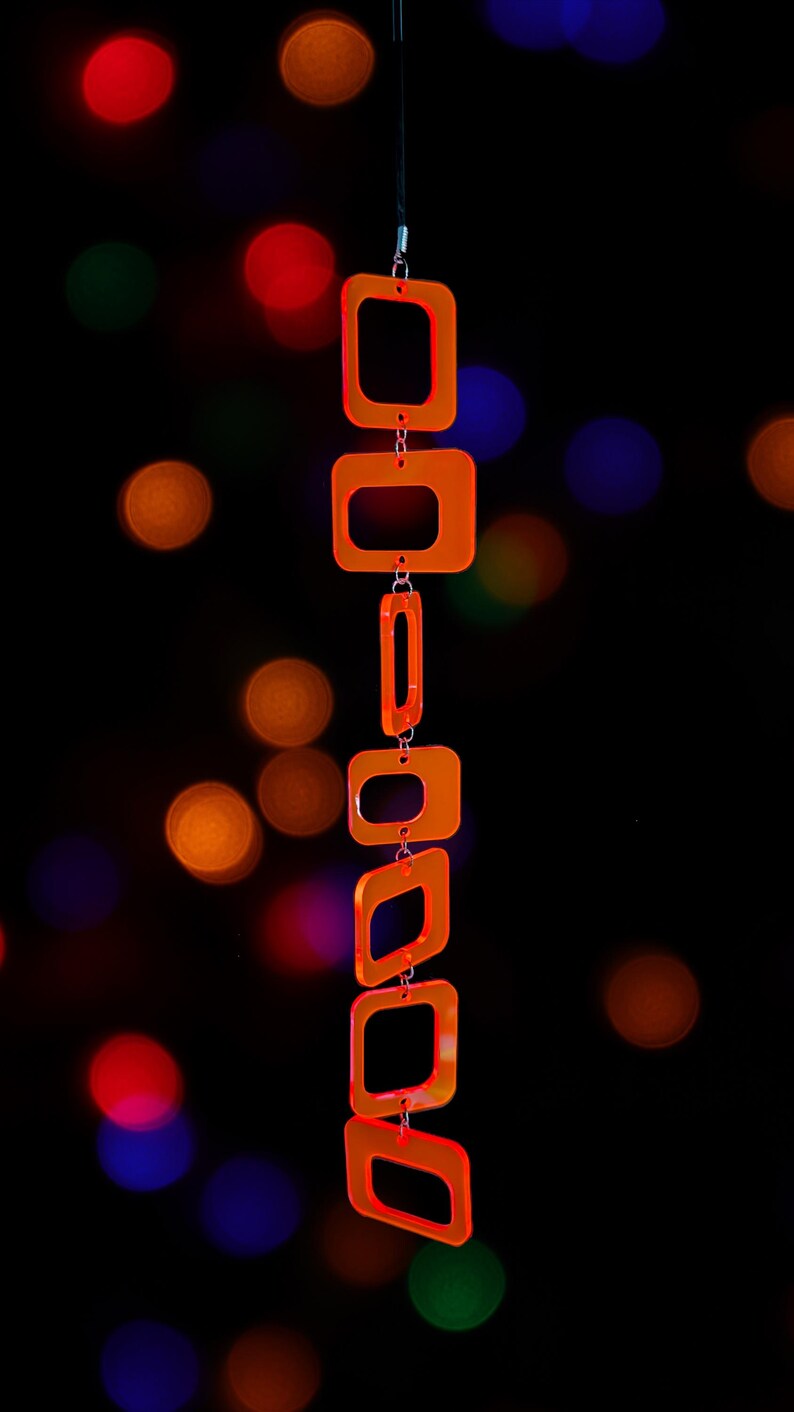 May include: A vertical hanging ornament composed of six translucent neon orange acrylic square and rectangular frames linked together by small silver metal jump rings, suspended from a thin black cord against a dark background.