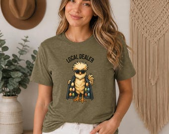 May include: An olive green short-sleeve t-shirt featuring a graphic of a yellow chick wearing sunglasses and an open coat. The chick is displaying various colored eggs inside the coat. The text above the chick reads LOCAL DEALER.
