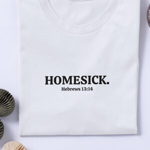 Op de afbeelding: Een wit t-shirt met het woord "HOMESICK." in het zwart gedrukt, met "Hebrews 13:14" eronder. Het shirt ligt plat op een wit oppervlak, met decoratieve papieren ornamenten en een kleine plant in een decoratieve pot.
