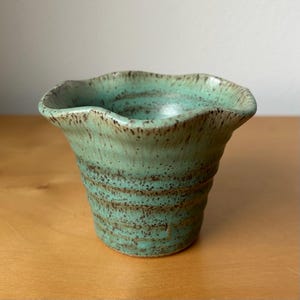 May include: A handmade ceramic vase with a unique, wavy rim. The vase features a turquoise glaze with brown speckles, giving it a rustic appearance. The cylindrical body has a textured, ribbed design.