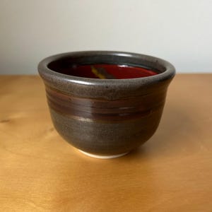 May include: A small ceramic bowl with a dark, textured exterior featuring horizontal bands of brown and charcoal gray. The interior of the bowl is finished with a smooth, glossy red glaze that contrasts with the muted tones of the outer surface.