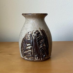 May include: A small, speckled beige ceramic vase with a flared rim and a wide, rounded body. The exterior features a prominent, dark brown, textured abstract relief design that wraps around the front, contrasting with the smooth, light clay surface.