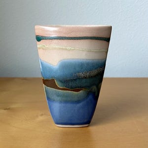 May include: A ceramic vase with a unique, layered design. The top features a soft pink hue, transitioning to bands of green, cream, and blue. A dark brown band separates the blue from the darker blue base. The vase has a slightly flared shape.