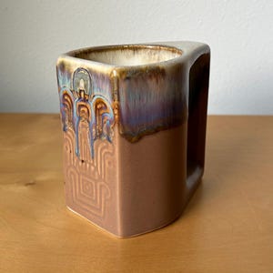 May include: A brown ceramic mug with a unique triangular shape and a handle. The mug features an embossed design of figures and geometric patterns. The rim has a glossy, iridescent glaze with shades of blue, purple, and brown.