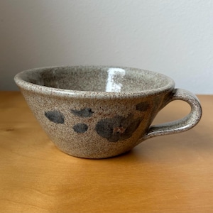 May include: A speckled ceramic mug with a rounded body and a handle. The mug is primarily tan with dark gray and brown floral-like patterns. The mug sits on a wooden surface.