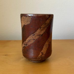 May include: A handmade ceramic cup with a dark reddish-brown glaze. Diagonal bands of lighter brown cross the cup's surface. The cup has a rounded base and a slightly flared rim, showcasing a rustic aesthetic.