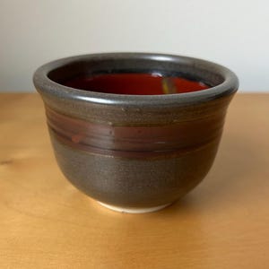 May include: A handmade ceramic bowl with a dark brown exterior and a vibrant red interior. The bowl features a horizontal band of a lighter brown color around the middle. The rim is a darker shade of brown. The bowl is likely used for serving food.