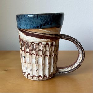May include: A ceramic mug featuring a dark blue glazed rim and a body with a marbled brown and white pattern. The lower section of the mug has vertical fluted grooves, and it includes a curved handle with a matching marbled brown and white finish.