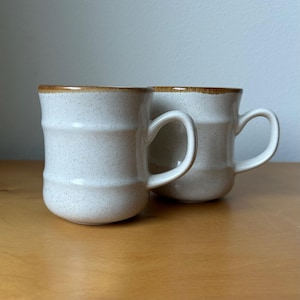 May include: Two off-white ceramic mugs with a speckled finish and a brown rim. The mugs have a cylindrical shape with a handle and are set on a wooden surface. The mugs are a neutral color and would be suitable for coffee or tea.