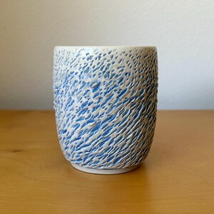 May include: A cylindrical ceramic cup featuring a textured exterior with a raised, white, organic pattern layered over a vibrant blue base. The vessel has a smooth, flat rim and a slightly tapered base, showcasing a distinct contrast between the blue and white colors across its entire surface.