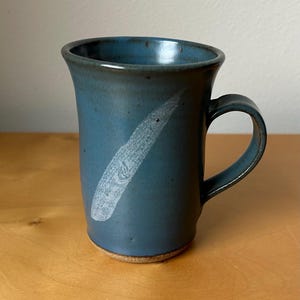 May include: A handmade ceramic mug in a muted teal color with a white, textured diagonal design. The mug has a rounded handle and a slightly flared rim. The base is a natural, unglazed clay color.