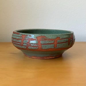 May include: A round ceramic bowl featuring a speckled sage green glaze with horizontal and vertical bands of exposed reddish-brown clay. The bowl has a wide, open top and a slightly tapered base, showcasing a textured finish on the exterior surface.