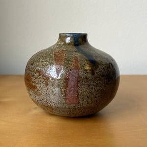 May include: A small, round ceramic vase with a narrow neck and a speckled, earthy texture. The surface features mottled shades of brown, gray, and tan, with distinct vertical streaks of dark blue and reddish-brown glaze accents across the body.