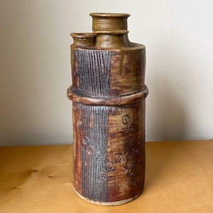 May include: A tall, handcrafted ceramic vase in shades of brown and tan. The vase features a cylindrical body with textured vertical lines and circular patterns. It has a unique design with a small spout and a wider opening at the top, showcasing a rustic aesthetic.