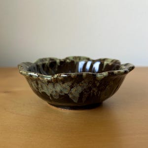 May include: A handmade ceramic bowl with a scalloped rim. The bowl features a dark brown glaze with green and tan accents. The bowl is round and sits on a small, circular base. The bowl is suitable for serving food.