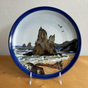 May include: A ceramic plate with a hand-painted coastal scene. The plate features a blue rim and a central image of a rocky coastline with a sandy beach, ocean waves, and two birds in flight. The plate is displayed on a clear stand.