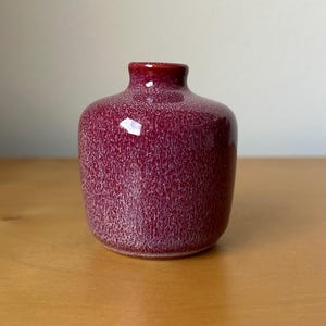 May include: A small, burgundy-colored ceramic vase with a speckled texture. The vase has a rounded body and a narrow neck. The surface has a glossy finish, reflecting light. The vase is sitting on a wooden surface.