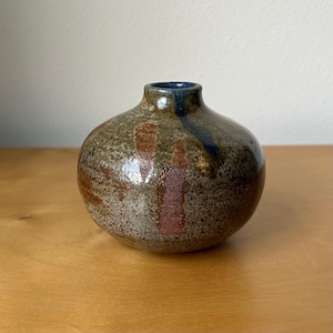 May include: A small, round ceramic vase with a speckled, earthy brown and gray glaze. The surface features vertical, dark reddish-brown brushstroke accents and a deep blue drip pattern extending from the narrow, circular opening at the top.