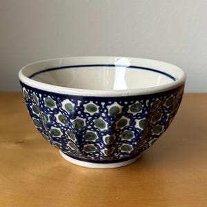 May include: A ceramic bowl with a cream-colored interior and a navy blue and green patterned exterior. The bowl has a scalloped edge and a dark blue rim. The pattern consists of green dots within a navy blue design.