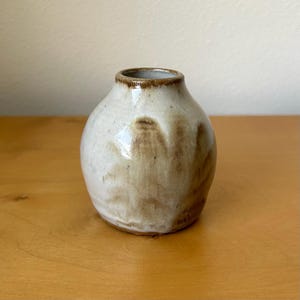May include: A small, round ceramic vase with a narrow neck and a wide, bulbous body. The piece features a glossy, off-white glaze with organic, flowing brown drip patterns that extend from the rim down the sides of the vessel.