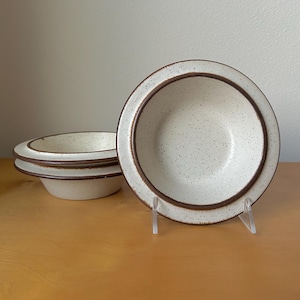 May include: A set of four ceramic bowls. The bowls have a speckled, off-white interior and a brown rim. The bowls are displayed on a wooden surface. The bowls are round and have a simple, classic design.
