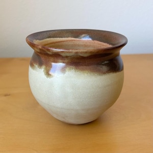 May include: A handmade ceramic pot with a rounded shape. The pot features a cream-colored base, transitioning to a band of brown and tan around the rim. The interior is a lighter tan color.