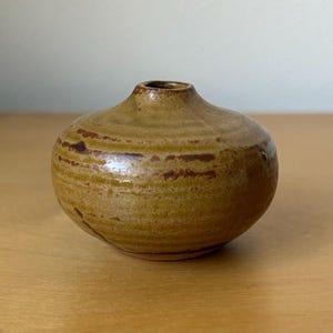 May include: A small, round ceramic vase with a narrow neck. The vase has a speckled, earthy brown and tan color scheme with darker brown horizontal stripes. The vase is sitting on a light brown wooden surface.