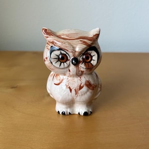 May include: A ceramic owl figurine with a marbled brown and cream design. The owl has large, detailed eyes with brown irises and black and white accents. The owl's feet are black, and the figurine is standing on a wooden surface.