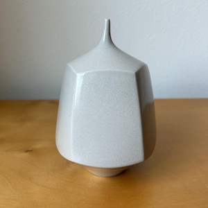 May include: A light gray ceramic vase with a faceted, geometric body and a narrow, tapered neck. The surface has a smooth, matte finish with subtle speckling throughout, featuring a distinct angular structure that tapers toward the top opening.