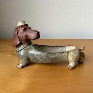 May include: A ceramic dachshund figurine with a brown head and a small hat. The dog-shaped container has a gray and beige body with a long, open trough. The figurine is a decorative item.