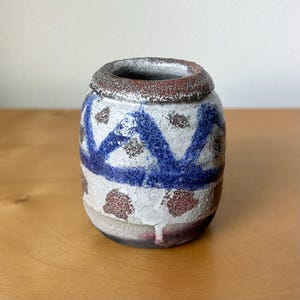 May include: A handcrafted ceramic vase with a cylindrical shape. The vase features a white base with blue geometric patterns and brown speckled accents. The rim is a dark red-brown color.