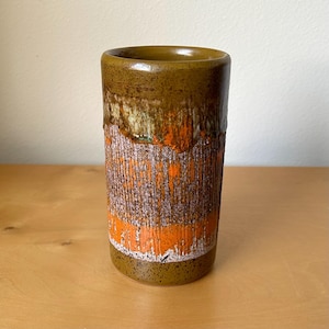 May include: A cylindrical ceramic vase featuring a smooth, dark olive-green rim and base. The central body displays a textured, vertical ribbed pattern with a layered glaze of vibrant orange, earthy brown, and off-white tones throughout.