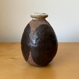 May include: A small, egg-shaped ceramic vase with a narrow neck and a flared rim. The body features a textured, dark brown glaze with exposed, rough, tan-colored clay sections. The rim is a smooth, light cream color with subtle brown speckles.