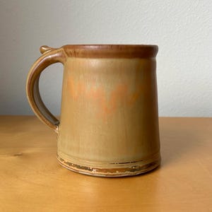 May include: A handmade ceramic mug with a handle. The mug is a light brown color with a darker brown rim and handle. There is an orange design on the front of the mug. The mug is sitting on a wooden surface.