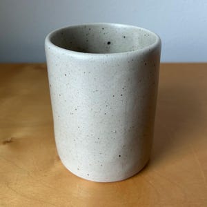 May include: A speckled, off-white ceramic tumbler with a rounded base and a smooth, cylindrical body. The interior is a darker shade of the same color. The cup is approximately 4 inches tall and is suitable for beverages.