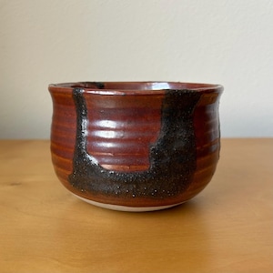 May include: A handmade ceramic bowl with a reddish-brown glaze and a dark, textured black accent. The bowl has a rounded shape and a slightly flared rim. The interior is a lighter shade of brown.