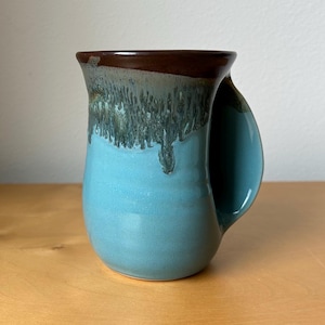 May include: A handmade ceramic mug with a unique design. The mug features a turquoise blue base, transitioning to a dark brown rim. The handle is a curved, open design. The glaze has a dripped effect, adding texture and visual interest to the piece.