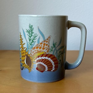 May include: A ceramic mug with a speckled off-white upper body and a light blue base. The exterior features a colorful graphic print of various seashells in shades of orange, brown, and yellow, accented by green and blue seaweed illustrations.